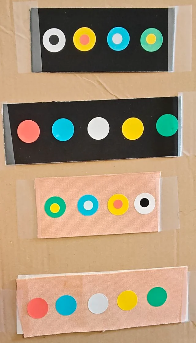 A cardboard board with four strips of kinesiology tape, two black and two beige. Each carries round stickers: plain red, pale blue, white, yellow and green, and dots in dots: white with a black middle, yellow with a red middle, blue with a white middle and green with a yellow middle.
